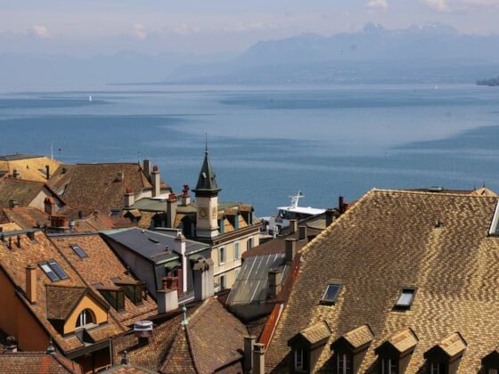 View over a European harbor town with terracotta rooftops toward a calm blue lake and distant mountains in the background with a church steeple nearby.
