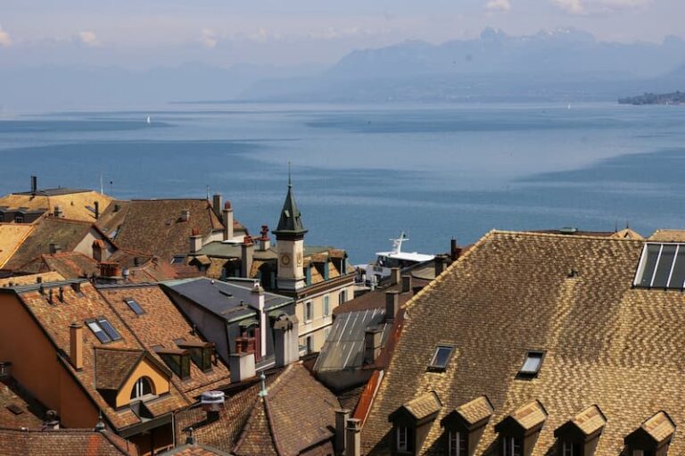 View over a European harbor town with terracotta rooftops toward a calm blue lake and distant mountains in the background with a church steeple nearby.