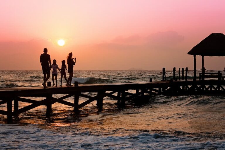 Silhouetted family walking hand in hand on a wooden pier at sunset over the ocean, pink sky above.