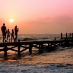 Silhouetted family walking on a wooden pier at sunset over the ocean edge.