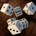 Five white dice with black pips tumble on a dark wooden surface, shadows visible underneath.