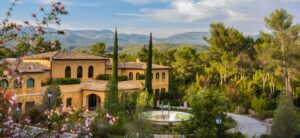 Sunny Mediterranean villa with yellow stucco walls, cypress trees, and a circular fountain in a landscaped courtyard.