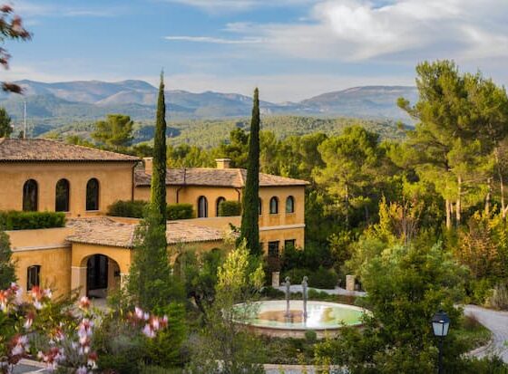 Sunny Mediterranean villa with yellow stucco walls, cypress trees, and a circular fountain in a landscaped courtyard.