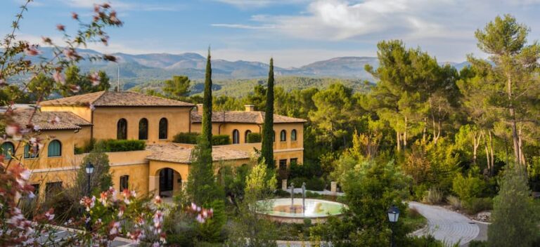Sunny Mediterranean villa with yellow stucco walls, cypress trees, and a circular fountain in a landscaped courtyard.