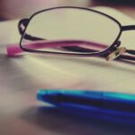 Close-up of black-rimmed glasses resting on lined notebook paper, with a blue pen in the foreground.