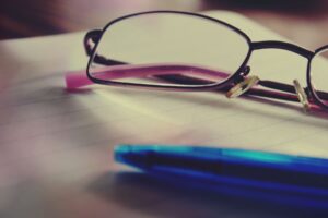 Close-up of black-rimmed glasses resting on lined notebook paper, with a blue pen in the foreground.