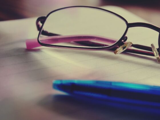 Close-up of black-rimmed glasses resting on lined notebook paper, with a blue pen in the foreground.
