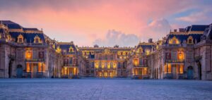 Grand palace courtyard at dusk, warm lights glow from ornate buildings surrounding a cobblestone square under a pink-orange sky.