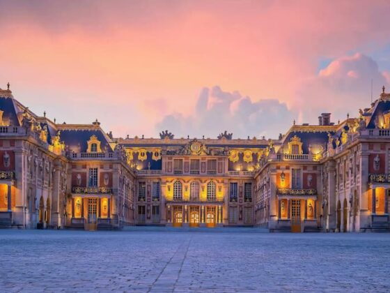 Grand palace courtyard at dusk, warm lights glow from ornate buildings surrounding a cobblestone square under a pink-orange sky.
